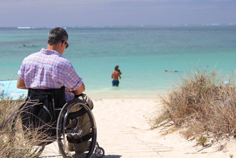 Mann im Rollstuhl in Sanddünen im Hintergrund blaues Meer